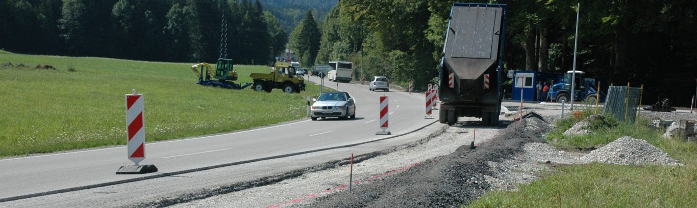 Bauarbeiten an einer Straße mit Verkehr und Absperrungen in grüner Landschaft.